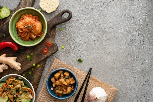 Vista dall'alto di ciotole con kimchi su taglieri vicino bacchette, aglio, zenzero e peperoncino sulla superficie di cemento — Foto stock