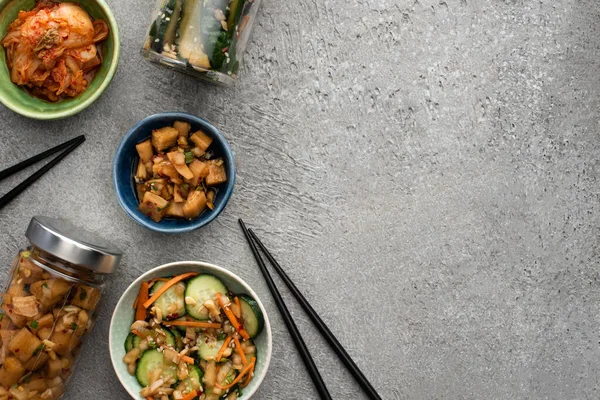 Vue de dessus des kimchi savoureux dans des bols et des bocaux près des baguettes sur la surface du béton — Photo de stock