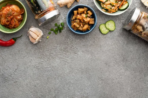 Blick von oben auf leckere Kimchi in Schalen und Gläsern in der Nähe von Essstäbchen, Knoblauch und Chilischote auf Betonoberfläche — Stockfoto