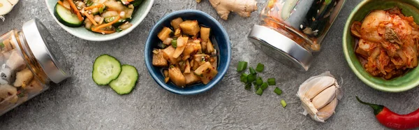 Panoramaaufnahme schmackhafter Kimchi in Schüsseln und Gläsern in der Nähe von Essstäbchen, Knoblauch und Chilischote auf Betonoberfläche — Stockfoto