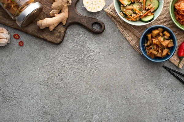 Blick von oben auf schmackhafte Kimchi in Schüsseln und Glas in der Nähe von Schneidebrett, Essstäbchen, Knoblauch und Ingwer auf Betonoberfläche — Stockfoto