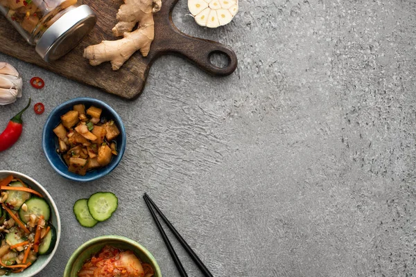 Vista dall'alto di gustosi kimchi in ciotole e vasetto vicino al tagliere, bacchette, aglio, zenzero e peperoncino sulla superficie di cemento — Foto stock