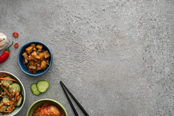 Top view of bowls with delicious kimchi near garlic, sliced cucumber, chili pepper and chopsticks on concrete surface — Stock Photo