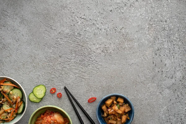 Vue de dessus des bols avec de délicieux kimchi près du concombre tranché et des baguettes sur la surface du béton — Photo de stock