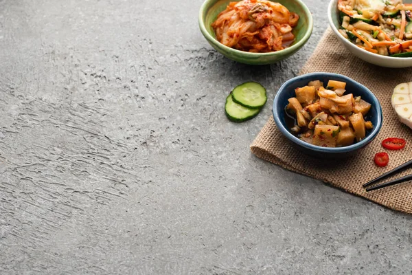 Bowls with kimchi on sackcloth near chopsticks, sliced garlic, cucumber and chili pepper on concrete surface — Stock Photo