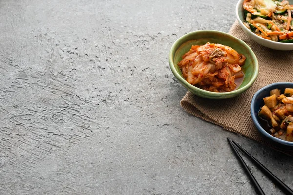 Bowls with kimchi on sackcloth near chopsticks on concrete surface — Stock Photo