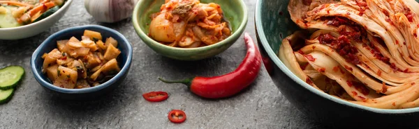 Panoramic shot of kimchi in bowls, chili pepper and garlic on concrete surface — Stock Photo