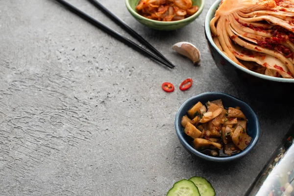 Bowls with delicious kimchi near sliced cucumber, garlic and chopsticks on concrete surface — Stock Photo