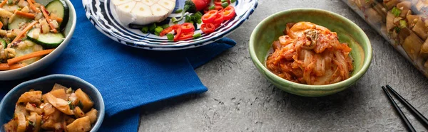 Plano panorámico de sabroso kimchi en cuencos y frasco en tela azul cerca de palillos en superficie de hormigón — Stock Photo