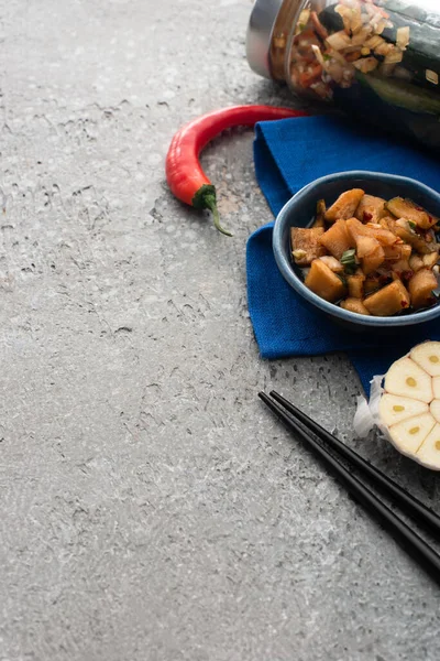 Sabroso kimchi en cuencos y frasco en tela azul cerca de palillos, chile y ajo en rodajas en la superficie de hormigón - foto de stock