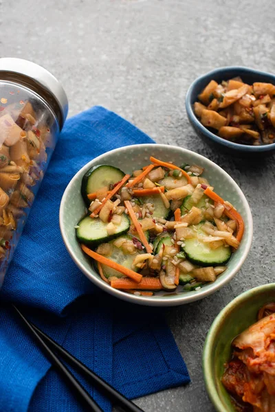 Deliciosos kimchi em tigelas e jarro perto de pauzinhos em pano azul na superfície de concreto — Fotografia de Stock