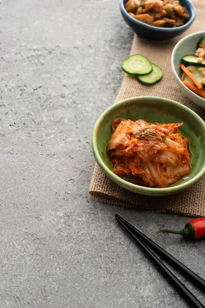 Taças de kimchi saboroso em pano de saco perto de pauzinhos na superfície de concreto — Fotografia de Stock