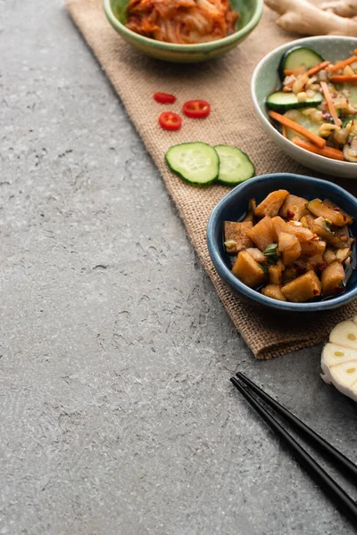 Selective focus of bowls with kimchi near ginger, sliced cucumber and chili pepper on sackcloth near chopsticks on concrete surface — Stock Photo