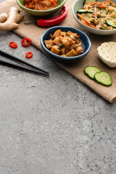 Taças de kimchi saboroso, pimenta, gengibre, alho fatiado e pepino em pano de saco perto de pauzinhos na superfície de concreto — Fotografia de Stock