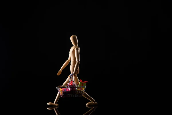 Wooden doll imitating walking with presents in shopping basket on black background — Stock Photo