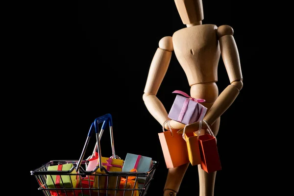 Boneca de madeira com sacos de compras e presentes em cesta de compras isolada em preto — Fotografia de Stock