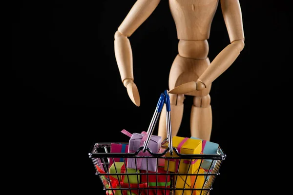 Foco seletivo de boneca de madeira e presentes na cesta de compras isolada em preto — Fotografia de Stock