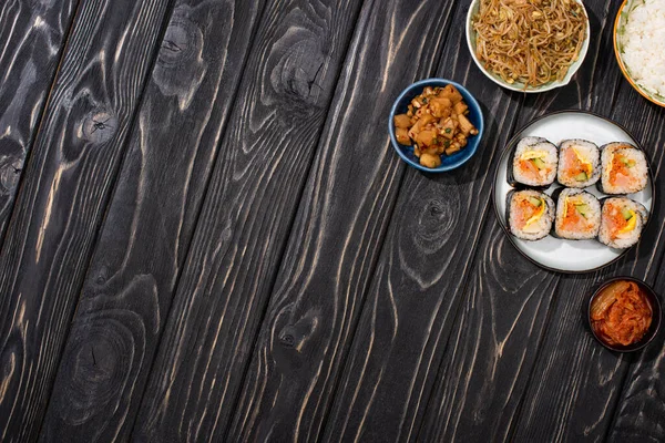 Top view of bowls with korean side dishes near tasty rice rolls on wooden surface — Stock Photo