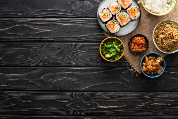 Draufsicht auf Schüsseln mit leckeren koreanischen Beilagen in der Nähe von Gimbap auf Holzoberfläche — Stockfoto