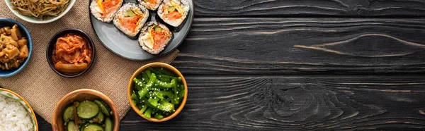Panoramic shot of tasty korean side dishes near rice rolls on wooden surface — Stock Photo