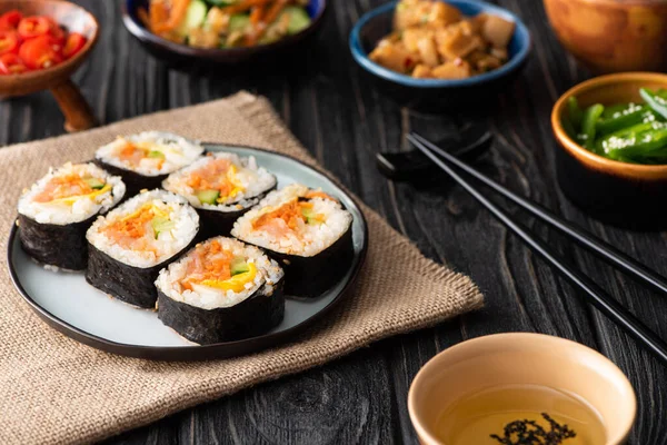 Messa a fuoco selettiva di gustosi gimbap con verdure e salmone vicino a bacchette e contorni su superficie di legno — Foto stock