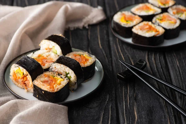 Selektiver Fokus von leckerem Gimbap mit Gemüse und Lachs auf Tellern in der Nähe von Essstäbchen auf Holzoberfläche — Stockfoto