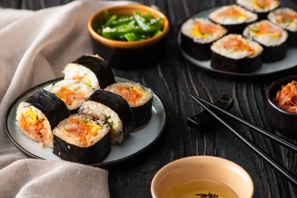 Selektiver Fokus schmackhafter Reisbrötchen auf Tellern in der Nähe von Essstäbchen und koreanischer Beilagen auf hölzerner Oberfläche — Stockfoto