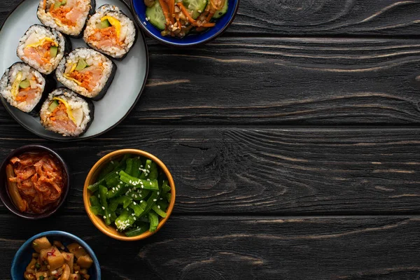 Vue de dessus des plats d'accompagnement coréens près de la plaque avec des rouleaux de riz sur la surface en bois — Photo de stock