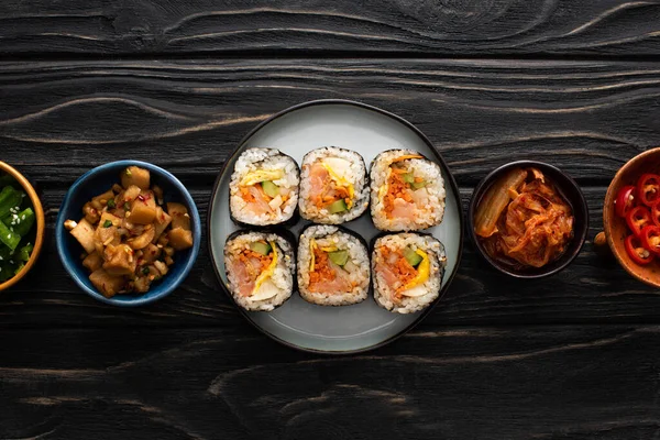 Teller mit Gimbap in der Nähe von Beilagen in Schüsseln auf Holzoberfläche — Stockfoto