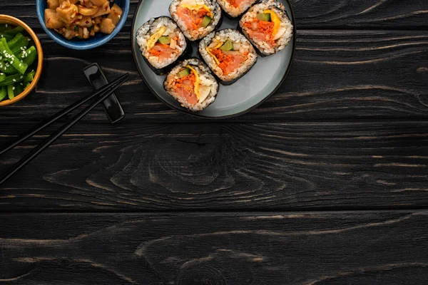 Vista superior de pauzinhos perto da placa com gimbap e pratos laterais coreanos na superfície de madeira — Fotografia de Stock