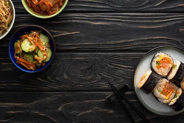 Верхний вид тарелки с корейским джимбапом и палочками рядом с вкусными гарнирами на деревянной поверхности — стоковое фото