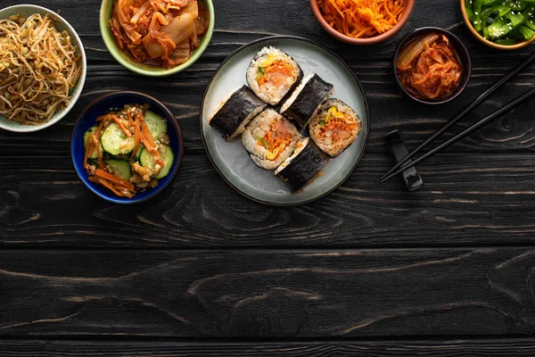 Верхний вид тарелки с гимбапом и палочками для еды рядом с вкусными корейскими гарнирами на деревянной поверхности — стоковое фото