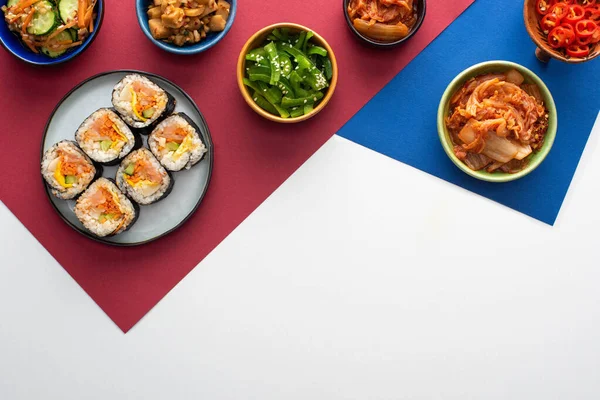 Верхний вид тарелки с вкусным джимбапом рядом с корейскими гарнирами на белом, синем и малиновом — стоковое фото