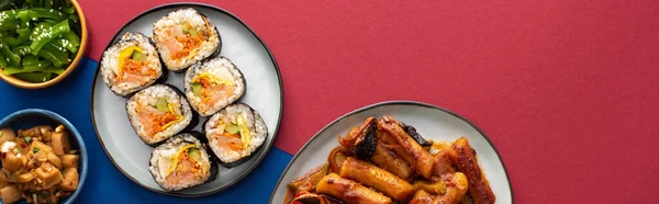 Panoramic shot of plates with tasty gimbap near korean side dishes on blue and crimson — Stock Photo