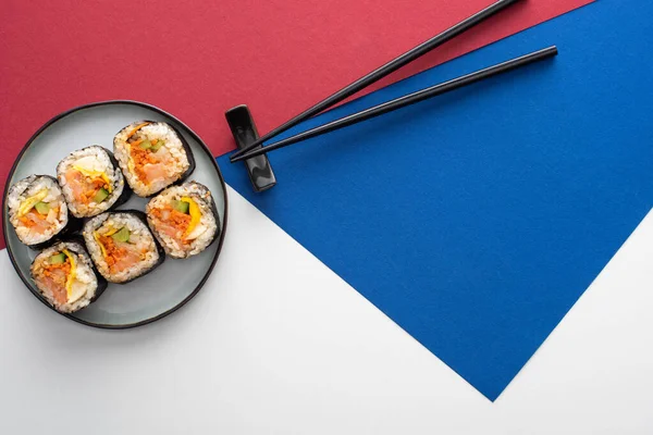 Vista dall'alto di bacchette vicino al piatto con gimbap su bianco, blu e cremisi — Foto stock