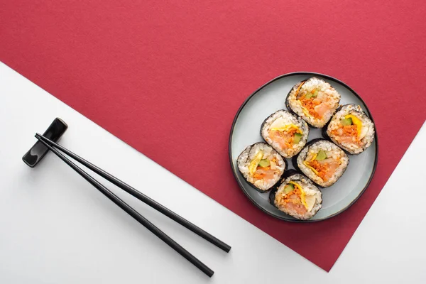 Vista dall'alto di bacchette vicino al piatto con gimbap su bianco e cremisi — Foto stock