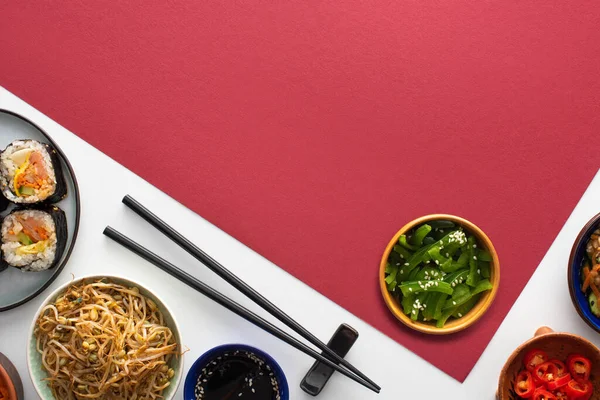 Vue de dessus du gimbap coréen frais et des plats d'accompagnement près des baguettes sur blanc et cramoisi — Photo de stock