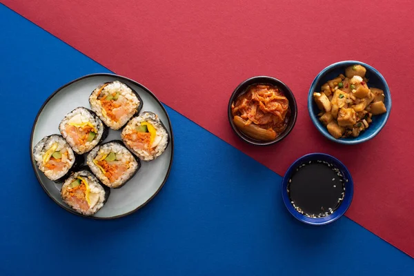 Vue du dessus de la sauce soja près de gimbap et des plats d'accompagnement coréens sur cramoisi et bleu — Photo de stock