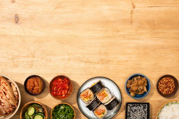 Top view of bowls with korean side dishes near korean gimbap on wooden surface — Stock Photo