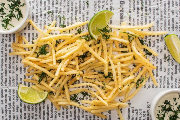 Vista superior de batatas fritas crocantes com endro perto de limão e molho de alho no jornal — Fotografia de Stock