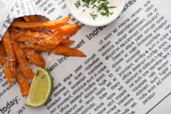 Vista superior de batatas fritas crocantes, limão fatiado e molho de alho com endro no jornal — Fotografia de Stock