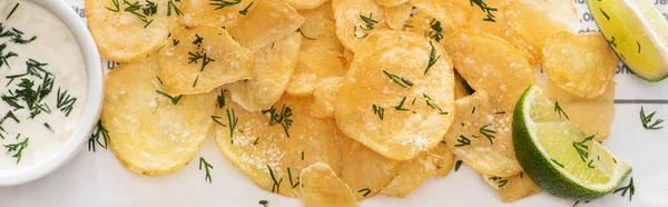 Панорамний знімок хрустких картопляних чіпсів з сіллю біля нарізаного лайма та часникового соусу на білому — стокове фото