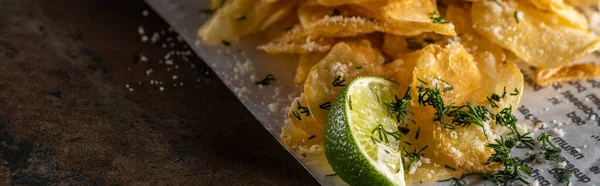 Panoramic shot of crunchy potato chips with salt near sliced lime and newspaper on marble surface — Stock Photo