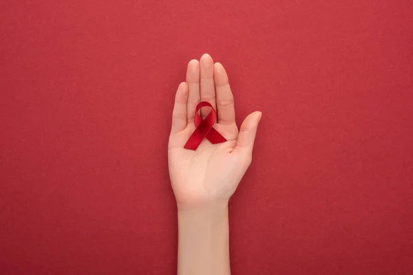 Cropped view of woman holding red awareness aids ribbon on red background — Stock Photo