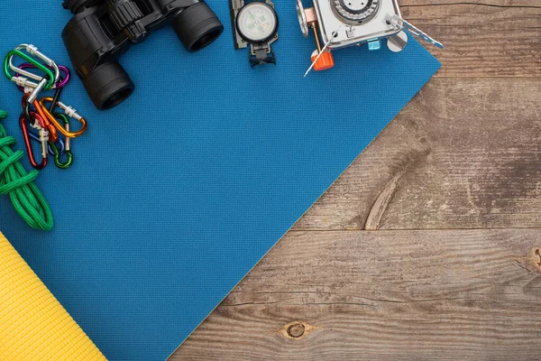 Top view of carabiners, hiking rope, binoculars, compass and gas burner on blue sleeping mat on wooden surface — Stock Photo