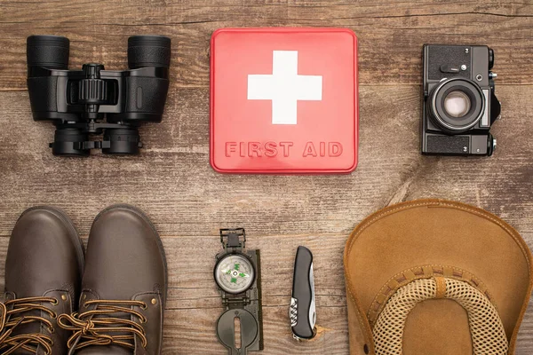 Vista dall'alto kit di pronto soccorso rosso, binocolo, macchina fotografica, stivali, cappello, bussola e jackknife su superficie di legno — Foto stock