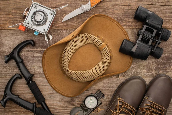 Vue de dessus du chapeau, des bottes, des bâtons de randonnée, des jumelles, du couteau à jackknife et du brûleur à gaz sur la surface en bois — Photo de stock