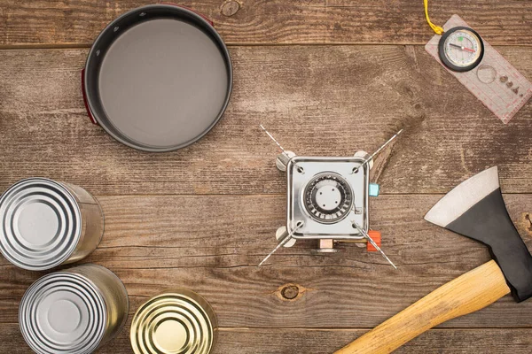 Top view of gas burner, axe, compass, metal bowl and canned goods on wooden surface — Stock Photo