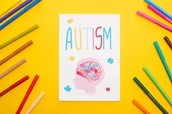 Visão superior de folha de papel com letras autismo e desenho de criança perto de canetas de feltro isoladas em amarelo — Fotografia de Stock