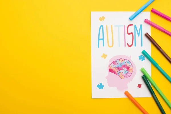 Vista dall'alto di pennarelli su foglio di carta con scritte autistiche e disegno di bambino isolato su giallo — Foto stock